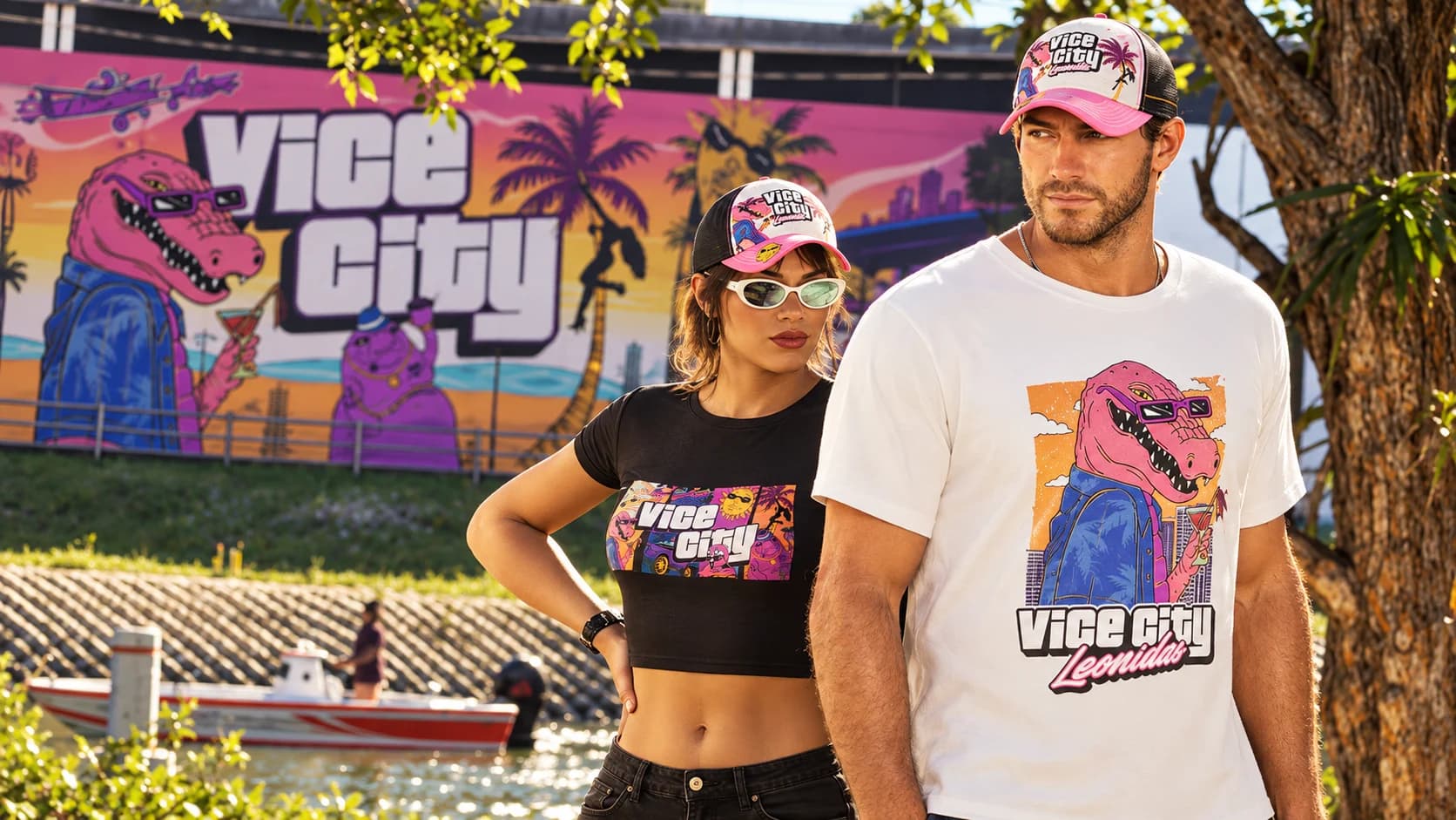 Two people wearing official Vice City Macca caps and tees in front of a giant Macca The Gator street mural by the water.
